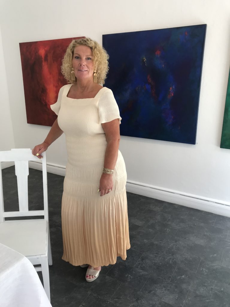 Ewa Kinnunen står i sitt galleri iklädd beigevit lång klänning och har ljust långt lockigt hår. Bakom syns två tavlor, en blå och en röd. 