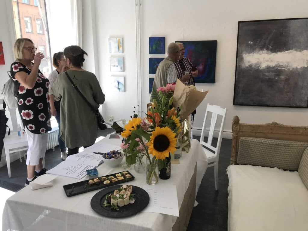 Besökare på versnissage med Ewa kinnunen. 5 personer syns i bild som står och pratar och i bakgrunden syns tavlor på väggen. I förgrunden finns ett bord med blommor och tilltugg på.